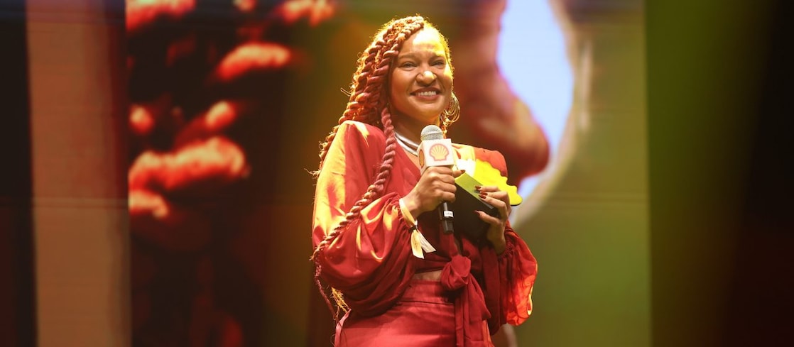 Pessoa em um palco segurando um microfone com a marca Shell e um troféu, sorrindo enquanto fala. Usa um vestido vermelho com mangas volumosas e cabelo trançado longo. Ao fundo, há iluminação em tons quentes e uma projeção ampliada de seu rosto.