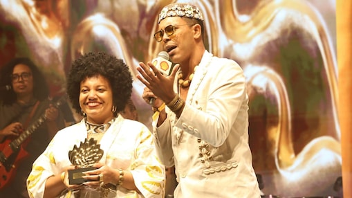 Duas pessoas estão no palco durante uma premiação, com iluminação quente e elementos dourados ao fundo. À esquerda, uma mulher sorridente com cabelo afro usa roupa branca com detalhes dourados e segura um troféu em formato de concha. À direita, um homem com óculos, colar e chapéu decorado fala ao microfone com a marca da Shell, enquanto gesticula com a mão.