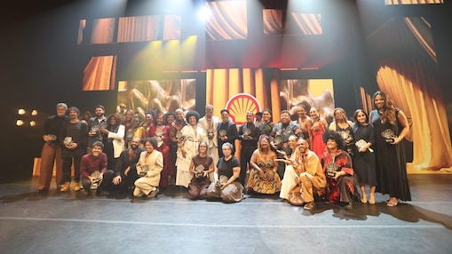Grupo grande de pessoas reunidas em um palco, posando para foto enquanto seguram troféus. Ao fundo, há iluminação cênica e telões com elementos visuais em tons dourados, além do logotipo da Shell ao centro. 