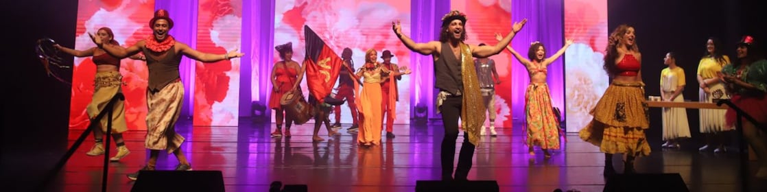 Elenco de artistas em trajes coloridos se apresenta no palco durante cerimônia do Prêmio Shell de Teatro, com painéis ao fundo exibindo flores vermelhas e iluminação cênica em tons de roxo.