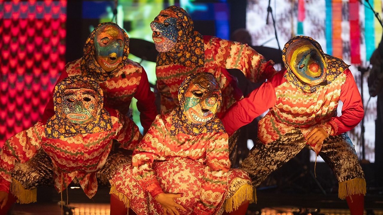 Artistas mascarados com figurinos floridos e vibrantes posam em cena durante apresentação teatral no Prêmio Shell de Teatro, em um momento performático que remete a manifestações populares brasileiras.