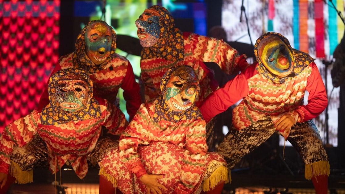 Artistas mascarados com figurinos floridos e vibrantes posam em cena durante apresentação teatral no Prêmio Shell de Teatro, em um momento performático que remete a manifestações populares brasileiras.