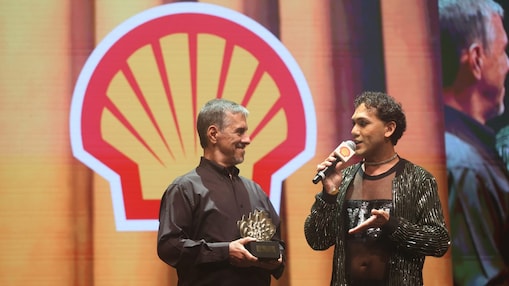 Duas pessoas estão no palco durante um evento, com o logotipo da Shell em grande destaque ao fundo. À esquerda, um homem de camisa preta segura um troféu em formato de concha e sorri. À direita, outra pessoa, vestindo um look preto com transparência e brilho, segura um microfone com a marca da Shell e fala enquanto gesticula.