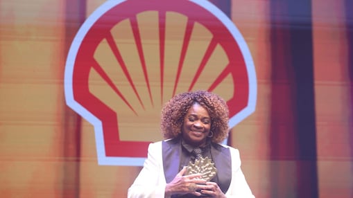 Pessoa em um palco segurando um troféu e sorrindo. Usa blazer branco com camisa escura e gravata. Ao fundo, há o logotipo da Shell em destaque e iluminação em tons quentes