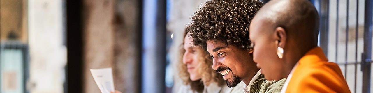 Três pessoas em um ambiente de trabalho colaborativo, sorrindo enquanto analisam documentos. Na mesa, há um laptop e copos de café.