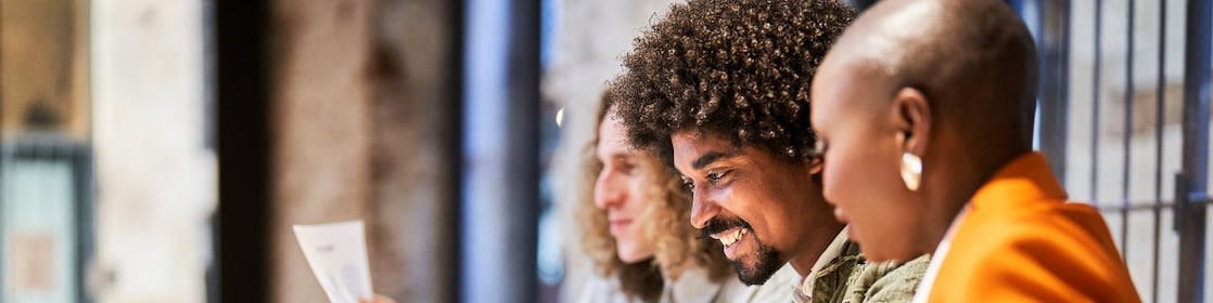 Três pessoas em um ambiente de trabalho colaborativo, sorrindo enquanto analisam documentos. Na mesa, há um laptop e copos de café.