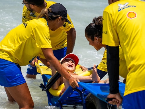 Um grupo de quatro voluntários com camisetas amarelas e bermudas azuis ajuda uma criança sorridente de boné vermelho, sentada em uma cadeira de rodas de praia adaptada, na beira da água. A cena demonstra trabalho em equipe e inclusão, enquanto eles ajudam a criança a aproveitar as ondas do mar.