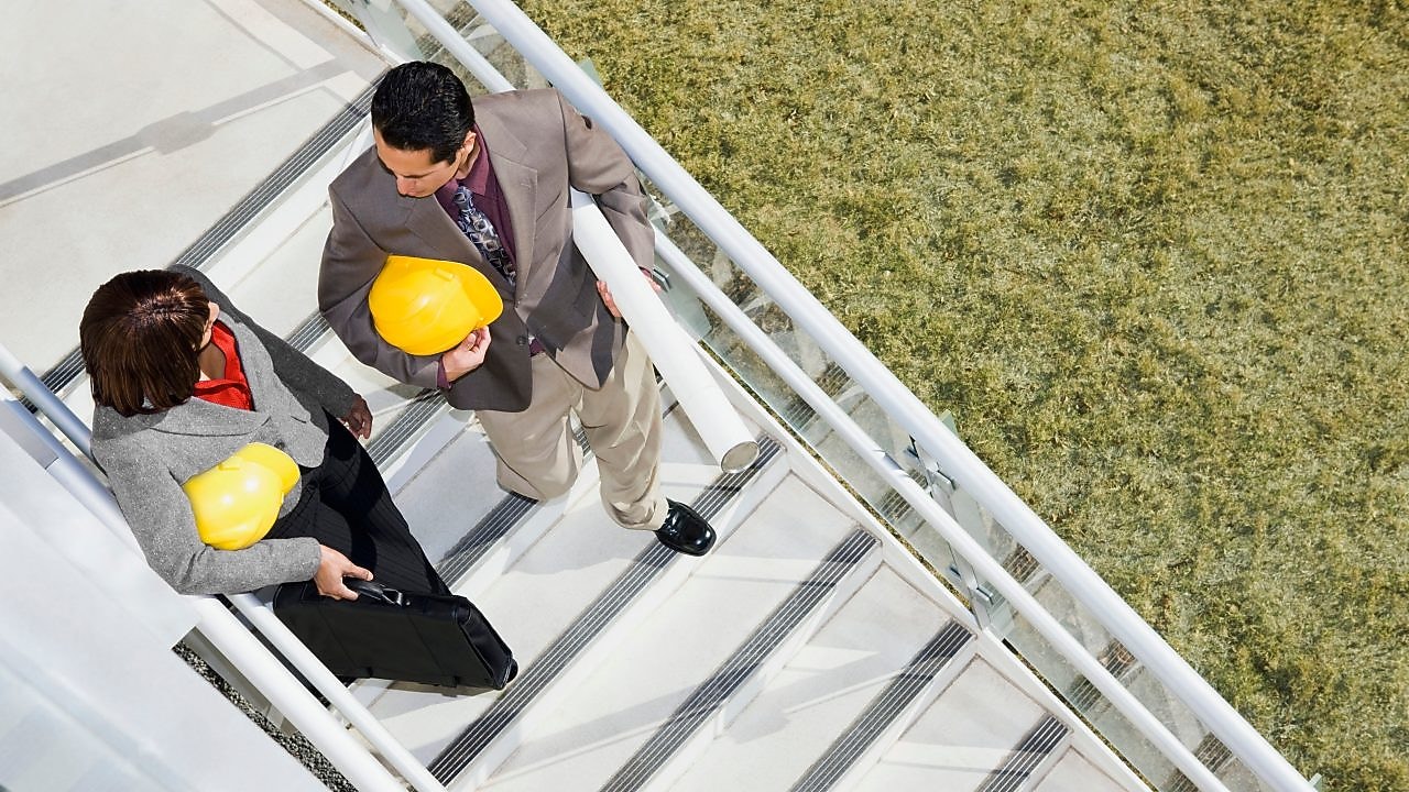 Funcionários, um homem e uma mulher, conversam entre si enquanto caminham numa escada e seguram capacetes de  trabalho