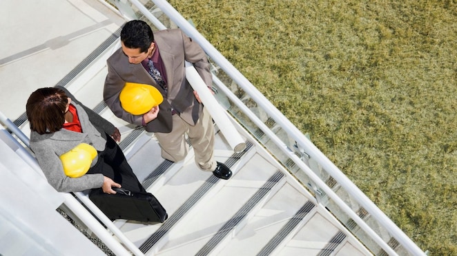 Funcionários, um homem e uma mulher, conversam entre si enquanto caminham numa escada e seguram capacetes de  trabalho