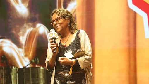 Mulher mais velha fala ao microfone em um palco iluminado em tons quentes, segurando um prêmio e um cartão, com o logotipo da Shell visível no microfone e uma imagem ampliada ao fundo.