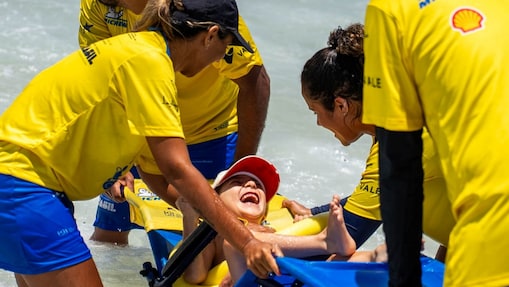 Quatro monitores usando camisetas amarelas e shorts azuis conduzem uma pessoa com deficiência em uma cadeira anfíbia até o mar. A pessoa está sorrindo amplamente, com a cabeça inclinada para trás, demonstrando alegria durante a experiência. Um dos monitores segura a parte frontal da cadeira enquanto outros a estabilizam pelas laterais e atrás. A cena acontece na beira da praia, com a água já envolvendo parte da cadeira, sugerindo uma atividade de acessibilidade e inclusão no ambiente marinho.